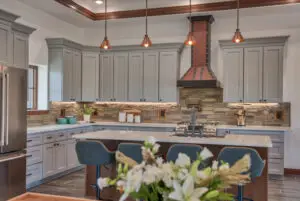 Interior photo of a home built by Harvell Homes in Powell, Wyoming featuring a luxurious custom kitchen with tray ceiling, premium appliances, wood island with premium countertop, wall-mount pot filler faucet, farmhouse kitchen sink, elevated stone backsplash, and kitchen hood