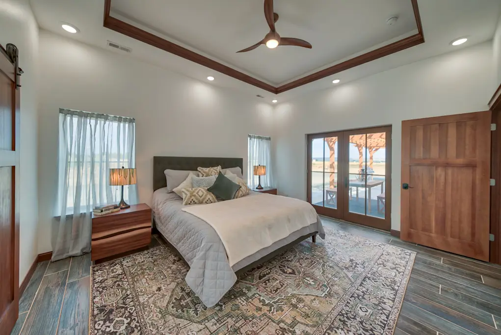 Interior photo of a home built by Harvell Homes in Powell, Wyoming featuring a luxurious and furnished bedroom with tray ceiling, balcony double french doors, and wood-look plank tile flooring